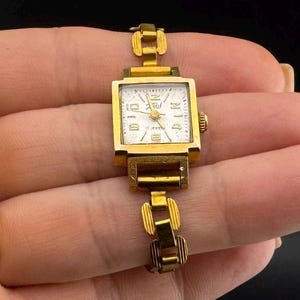 Könnte beinhalten: Eine Vintage-Armbanduhr in Goldoptik mit quadratischem Zifferblatt und goldenem Gliederarmband. Das Zifferblatt ist weiß mit schwarzen Ziffern und Zeigern. Die Uhr wird in einer Hand vor dunklem Hintergrund gehalten.