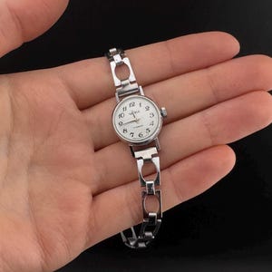 Könnte beinhalten: Eine silberfarbene Vintage-Armbanduhr mit weißem Zifferblatt und schwarzen Ziffern. Die Uhr hat ein Gliederarmband und eine kleine Krone. Auf dem Zifferblatt steht "Zaria" und "CCCP".