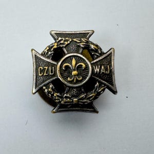 May include: An antique cross-shaped medal with the words "CZU" and "WAJ" inscribed on the arms. The centre features a fleur-de-lis emblem, encircled by a laurel wreath. The medal has a tarnished, aged appearance.
