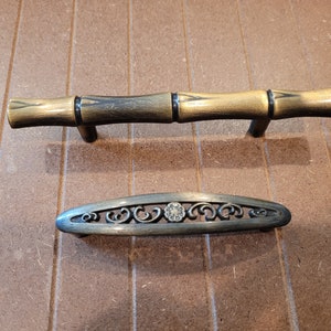 May include: Two antique-style cabinet door pulls. The top pull is a gold-toned bamboo design. The bottom pull is a silver-toned oval with a decorative floral design.
