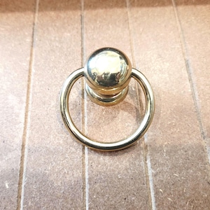 May include: A gold-colored ring pull with a spherical top and a circular handle. The hardware is made of metal and is designed for use on drawers, cabinets, or other furniture. The pull is shown against a light brown wooden surface.