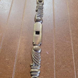 May include: A brass door latch with a decorative design. The latch has a square keyhole and a decorative design on the top and bottom.