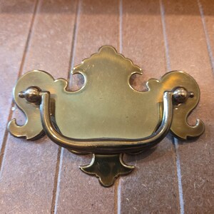 May include: A brass drawer pull featuring a decorative backplate with curved, symmetrical edges and a central shield-like shape. A U-shaped swinging handle is attached to the backplate by two circular mounts, allowing the handle to hang downward.