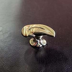 May include: A gold-toned cabinet knob with a unique, abstract design. The knob has a curved, wave-like shape and a smooth, polished finish.