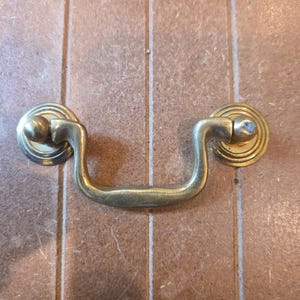 May include: A brass-colored metal drawer pull featuring a curved, U-shaped handle attached to two circular, tiered mounting plates. The hardware has a smooth, metallic finish with a slight patina and rounded ends on the handle attachment points.