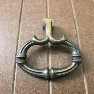 May include: Antique brass-toned drawer pull with an ornate design. The pull features a rectangular top with a curved handle and decorative detailing. Ideal for vintage furniture restoration projects.