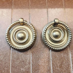 May include: Two antique brass drawer pulls. Each pull is circular with a central raised disc, surrounded by concentric rings and a beaded edge. A small, arched handle is attached at the top. The pulls have a warm, aged patina.