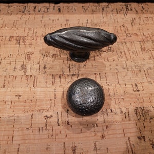 May include: Two black metal cabinet knobs. One is a round knob with a textured surface. The other is an oval knob with a twisted design.