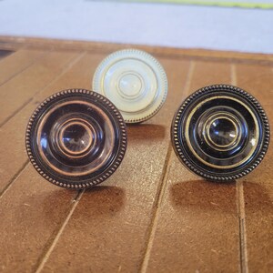 May include: Three decorative cabinet knobs in a row. One is dark brown, one is off-white, and the other is black with gold accents. Each knob has a circular design with a beaded edge, perfect for adding a touch of elegance to furniture.