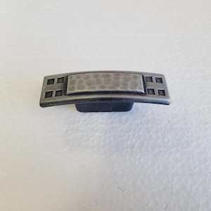 May include: A silver-toned cabinet pull with a hammered finish and a rectangular shape. The pull has a square design on each end.