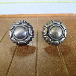 May include: Two antique-style cabinet knobs with a circular design and a metallic finish. The knobs have a central, raised circle surrounded by a decorative, scalloped edge. They are set against a brown wooden surface.