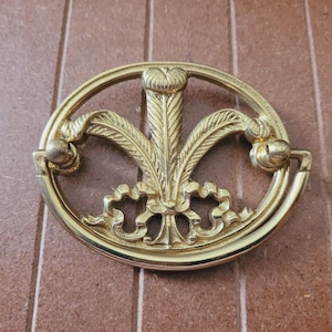 May include: A gold-toned metal cabinet pull with a decorative design featuring a stylized flower with leaves. The pull has a circular shape with a ring handle.