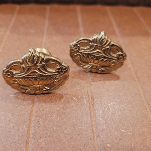 May include: Two antique brass cabinet knobs with ornate floral designs. The knobs have a gold finish and are oval-shaped with a raised, detailed pattern. They are likely vintage or reproduction hardware.