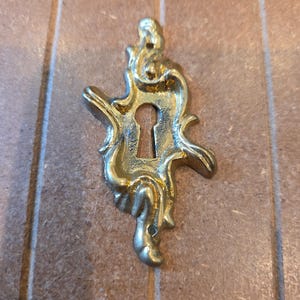 May include: A gold-colored metal keyhole escutcheon plate featuring an ornate, asymmetrical, swirling design. The piece has a central vertical keyhole opening and two small circular holes at the top and bottom for mounting purposes.