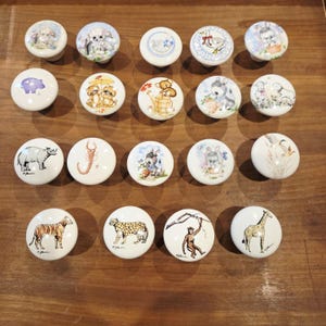 May include: A collection of twenty white ceramic cabinet knobs with various animal illustrations. The knobs feature images of a rhino, tiger, giraffe, and other animals. Each knob has a rounded shape and a metal screw.