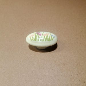 May include: A white ceramic oval-shaped cabinet knob with a floral design. The knob features a row of green leaves and pink, purple, and blue flowers.