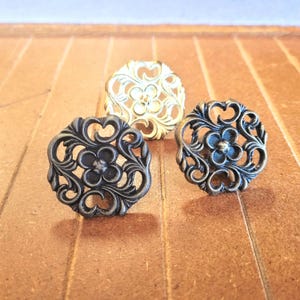 May include: Three decorative cabinet knobs with floral designs. Two knobs have a dark bronze finish, while the third is gold. Each knob features an intricate, openwork pattern with a central flower and swirling details.