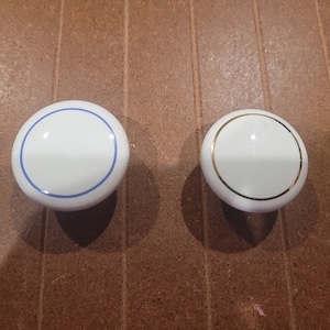 May include: Two white ceramic knobs with a single blue and gold trim around the edge.