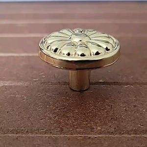 May include: A gold-colored cabinet knob with a floral design on the top. The knob has a round shape with a detailed pattern and a short, cylindrical stem. The knob is set against a brown wooden surface.