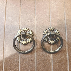 May include: Two antique brass cabinet pulls. Each pull features a decorative floral backplate with a ring handle. The pulls are mounted on a light brown wooden surface. The ring handles have a textured design.