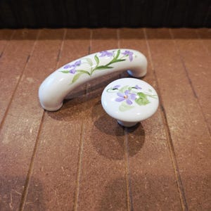May include: White ceramic cabinet hardware set featuring a curved handle and a round knob, both adorned with hand-painted purple violets and green leaves. The floral design adds a touch of elegance to any cabinet or drawer.