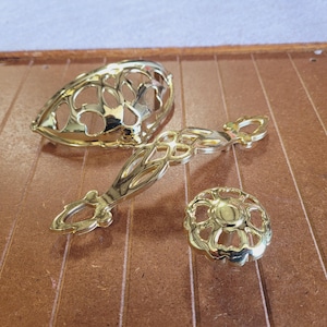 May include: Three gold-colored decorative hardware pieces. The set includes a handle, a knob, and a decorative oval-shaped piece. The hardware has an openwork design with floral and geometric patterns. The pieces are made of metal and are likely for furniture.