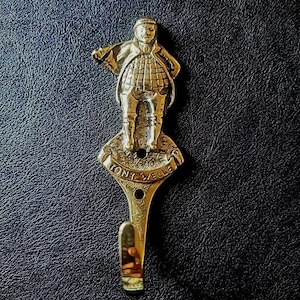 May include: A brass-coloured wall hook with a detailed figure of a man in traditional attire. The hook has the text "TONY VELLE" and is mounted on a dark, textured surface. The hook is designed for hanging items.