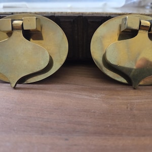 May include: Two antique brass drawer pulls. Each pull features a circular backplate with a stylized, teardrop-shaped handle. The pulls have a warm, golden hue and are mounted on a wooden surface.