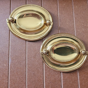 May include: Two gold oval cabinet knobs with a raised rim and a smaller oval inset. The knobs have a smooth, polished finish.