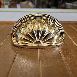 May include: A gold-colored, half-moon shaped cabinet pull with a shell-like design. The hardware has a polished finish and is set against a brown wooden surface. The pull is likely for a drawer or cabinet door.