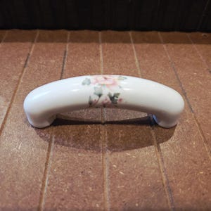 May include: White ceramic cabinet pull with a curved design. The pull features a floral pattern with pink and green accents. The hardware is designed for easy installation and adds a decorative touch to furniture.
