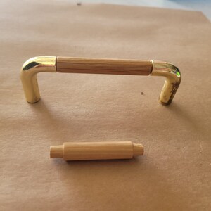 May include: A cabinet pull with a rectangular wooden center and curved, gold-colored metal ends. A separate, smaller wooden dowel is also present. The hardware is designed for furniture and home decor projects.