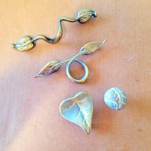 May include: Four silver leaf-shaped cabinet knobs. Two are long and curved, one is a ring with a leaf on each side, and one is a heart-shaped leaf.
