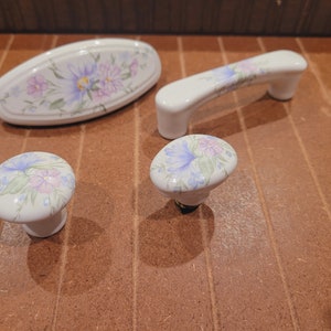 May include: Four ceramic drawer pulls with floral designs. The pulls are white with blue and pink flowers. Two of the pulls are round and two are oval.