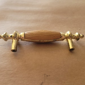 May include: A gold-coloured cabinet pull with a light brown wooden centre. The pull has a classic design with decorative end pieces and a cylindrical wooden handle. Suitable for furniture restoration or new builds.