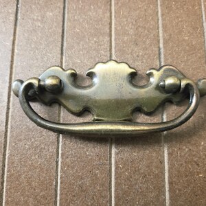 May include: Antique brass-toned drawer pull with a curved handle and decorative backplate. The hardware features a detailed, ornate design, perfect for vintage furniture restoration projects. The pull is approximately 5 inches wide.