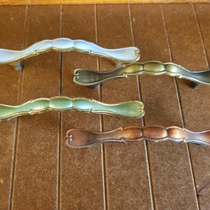 May include: Four decorative cabinet pulls in various colors: light blue, bronze, green, and copper. Each pull has a curved design with gold-colored accents, suitable for furniture restoration or DIY projects.
