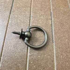 May include: A bronze-colored metal ring pull with a screw. The ring is circular and attached to a decorative base with a small ball detail. The screw is pointed and has a flat head.