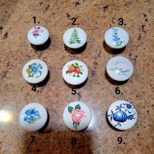 May include: Nine white ceramic cabinet knobs with floral designs. The knobs feature various colorful flower patterns, including pink roses, blue and pink floral arrangements, and a fern. Each knob is approximately 1.5 inches in diameter.