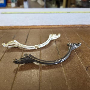 May include: Two decorative cabinet pulls with a curved design. One is a light gold color, and the other is a dark, matte gray. Both have ornate, floral-inspired details.