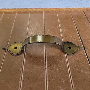 May include: A rustic, antique-style, metal door handle with a decorative, scalloped edge. The handle is a dark, aged brass color and has a simple, rounded shape.