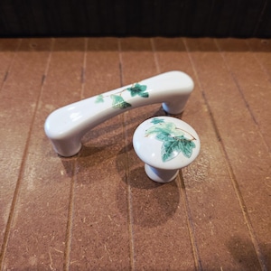 May include: White ceramic cabinet hardware set, featuring a handle and a knob. Both pieces are adorned with a green and blue ivy leaf design. The handle is approximately 5 inches long, and the knob is round.