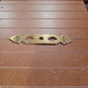 May include: A gold-colored metal decorative hardware piece with a rectangular shape and intricate cutouts. The piece has a square hole in the center and two smaller, oval-shaped cutouts on either side.