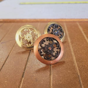 May include: Three decorative cabinet knobs in gold, copper, and black. Each knob features an ornate, openwork design with a floral center. The knobs are round and appear to be made of metal.