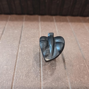 May include: A dark, carved wooden heart-shaped object with a stylized bird or angel design. The object is a deep black color and has a smooth, polished finish. The background is a light brown wooden surface.
