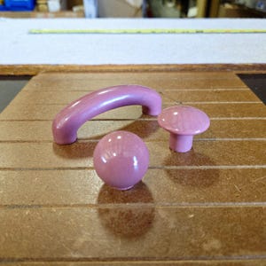 May include: A set of pink cabinet hardware, including a curved handle, a round knob, and a mushroom-shaped knob. The hardware is made of a glossy material and is set against a brown wooden surface.