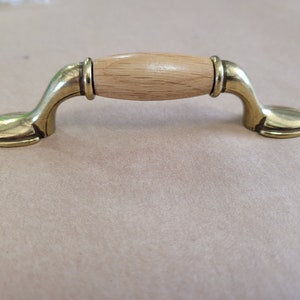 May include: A cabinet pull with a light wood centre and brass-coloured curved handles. The wooden centre is cylindrical, and the brass handles have a curved design with a rounded base. This hardware is suitable for cabinets and drawers.