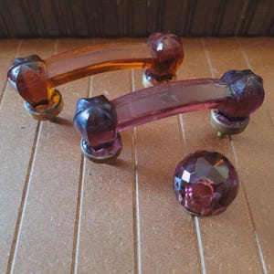 May include: Two vintage glass cabinet door pulls with a curved shape and faceted ends. One pull is amber glass and the other is purple glass. A single purple glass knob is also included.