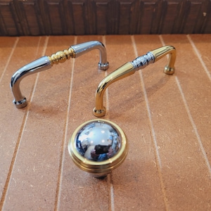 May include: Two gold cabinet pulls with a decorative design and a round, gold knob with a mirrored surface.