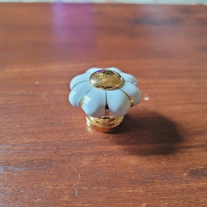 Könnte beinhalten: Weißer Keramik-Schrankknopf mit goldener Mitte und goldener Einfassung. Der Knopf ist blumenförmig mit fünf Blütenblättern.